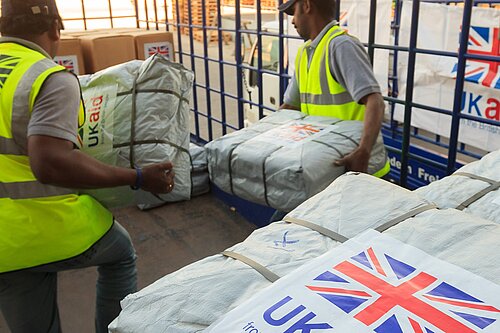 Two men are moving parcels wrapped in white plastic. In the foreground, a label on one package is visible reading "UK Aid from the British people" and a union flag. Image - Dfid CCBY-ND2.0